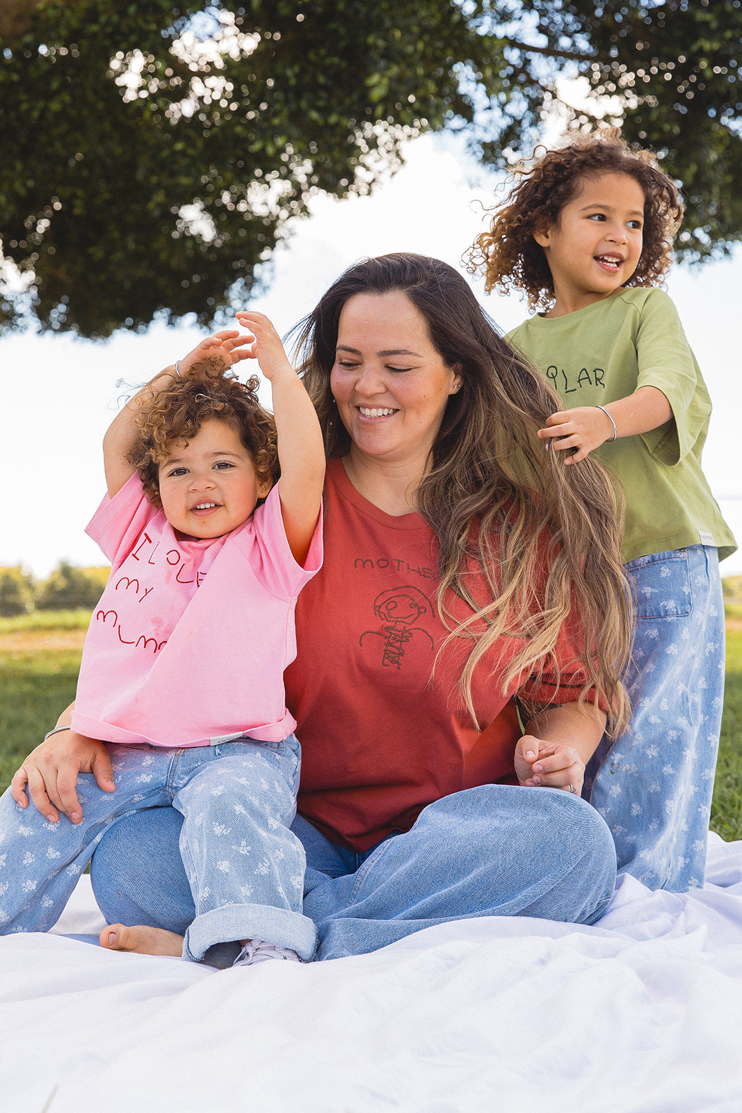 Momenta farm shoot: an adult in a red embroidered tee sitting outdoors with two children in pink and green embroidered tees.
