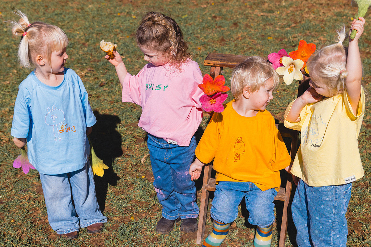 Momenta farm shoot: four children in blue, pink, orange and yellow tees with embroidered drawings and handwriting, holding colourful felt flowers.