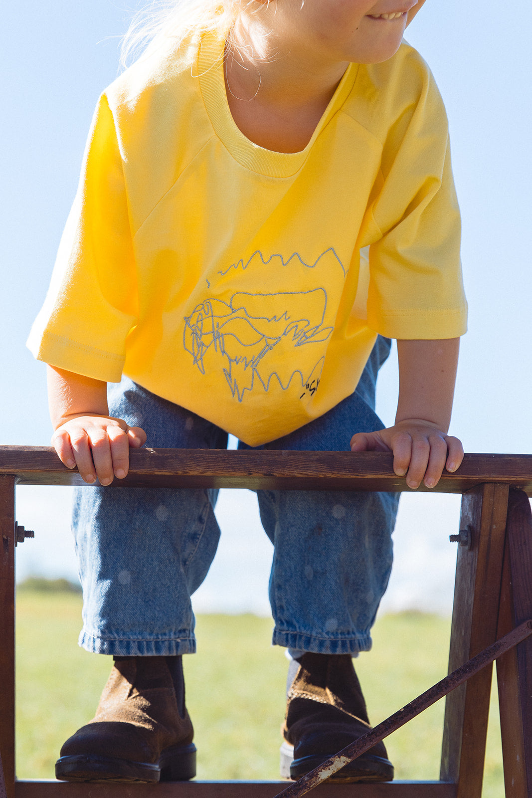 Momenta farm shoot: close-up of a child leaning on a wooden chair in a yellow tee with a grey embroidered line drawing.