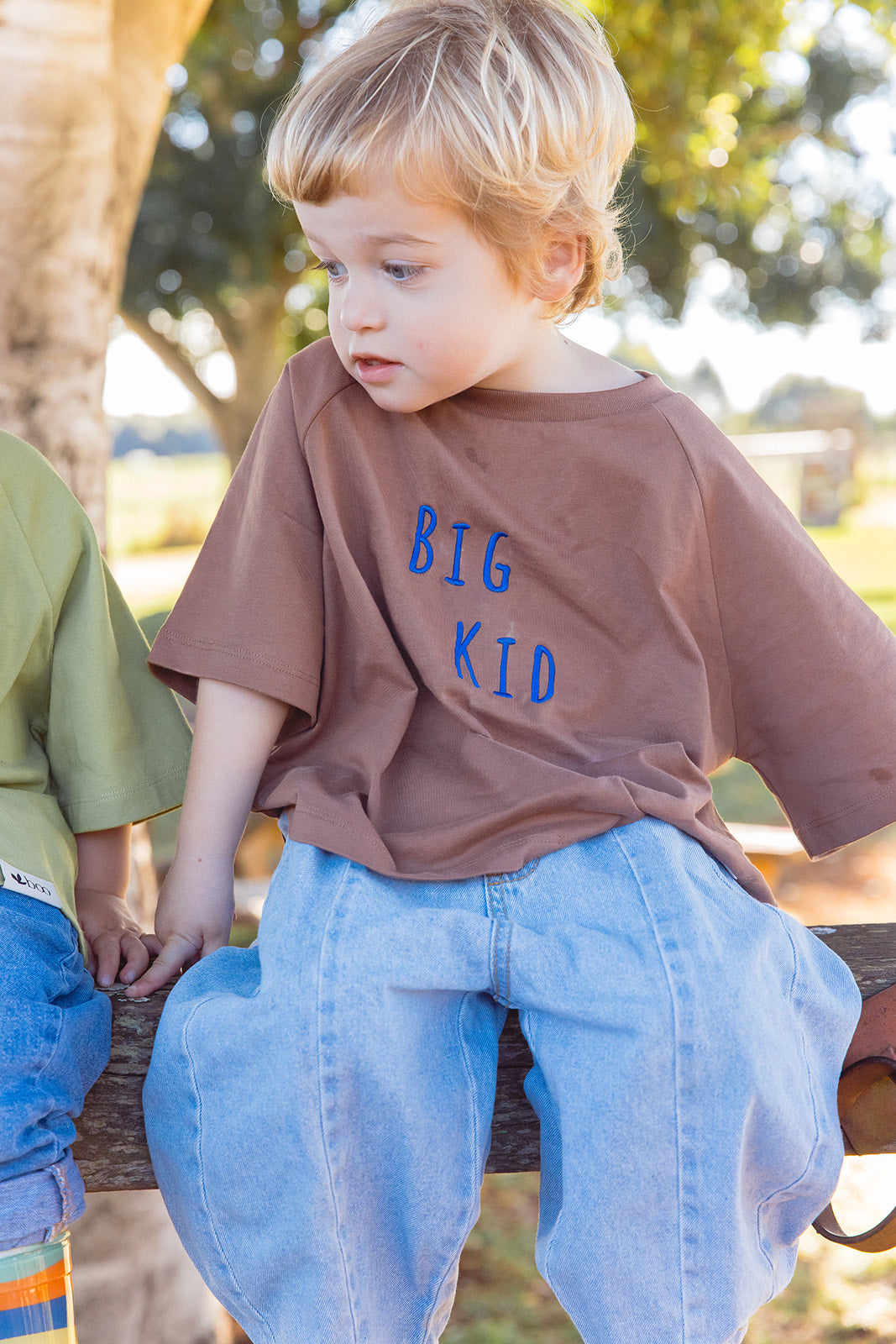 Momenta farm shoot: a child in a brown tee with blue BIG KID embroidery, seated on a wooden fence.